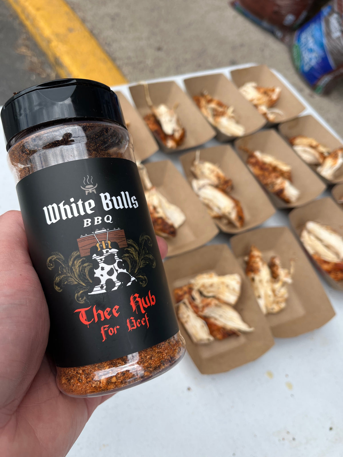 A hand holds a bottle of White Bulls BBQ seasoning "Thee Rub for Beef" in front of several small paper food trays containing cooked meat pieces on a white surface.
