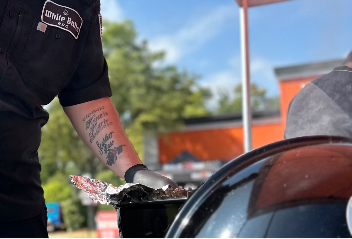 A man wearing a black branded short-sleeve shirt and black pants stands outdoors under an orange canopy next to a black kettle grill, preparing food.