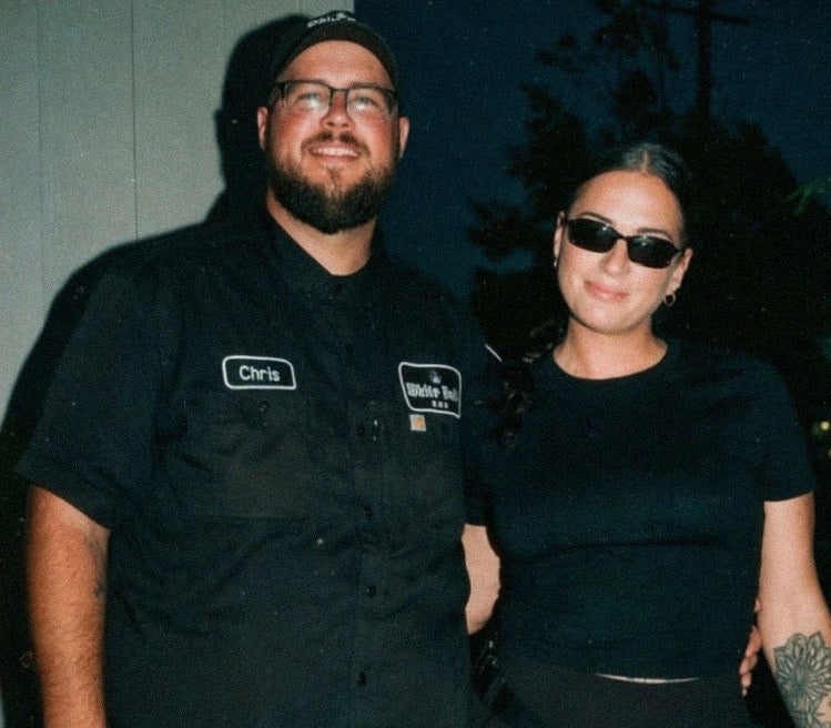 A man and a woman posing together at night. The man is wearing a black short-sleeve button-up shirt with a name tag reading 'Chris' and a patch that says 'Wicked', along with a black cap and glasses.