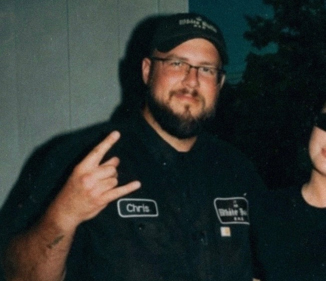 A man wearing a black button-up shirt with a name patch reading 'Chris' and a logo patch on the chest, standing outdoors at night and making a hand gesture. Another person is partially visible on the right.