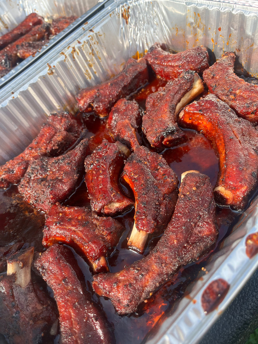 Close-up of glazed barbecue ribs coated in a dark reddish-brown sauce, arranged in a silver aluminum tray.