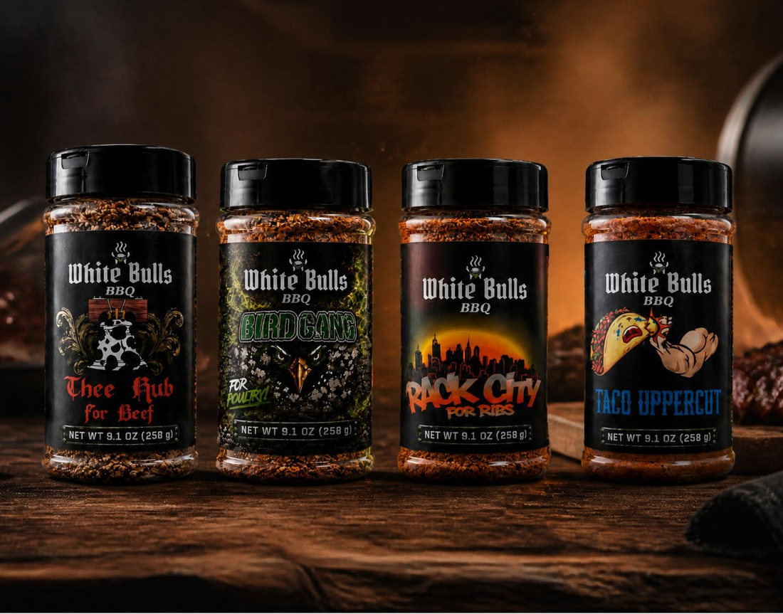 Four bottles of White Bulls BBQ seasoning blends arranged on a rustic wooden surface with cooked meat and cooking props in the background. The labels read Three Hub BBQ Salt, Nerd Food BBQ Rub, Rack City Rub, and Taco Uppercott BBQ Rub.