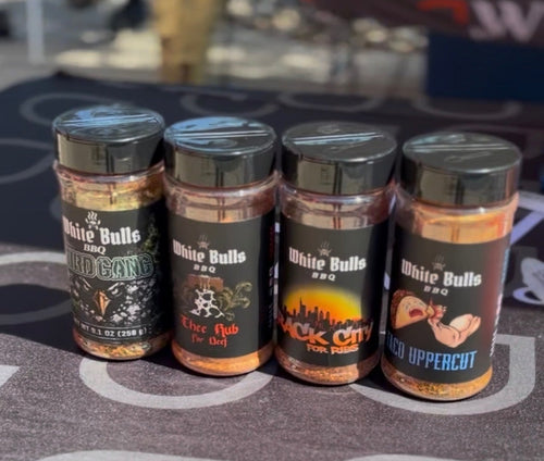 Four bottles of White Balls BBQ seasoning arranged on a branded table at an outdoor market or event. The bottles contain orange-brown spices and have black lids, with labels showing different flavor options: BBQ Rub, Smoke City, and Pepper Jack.